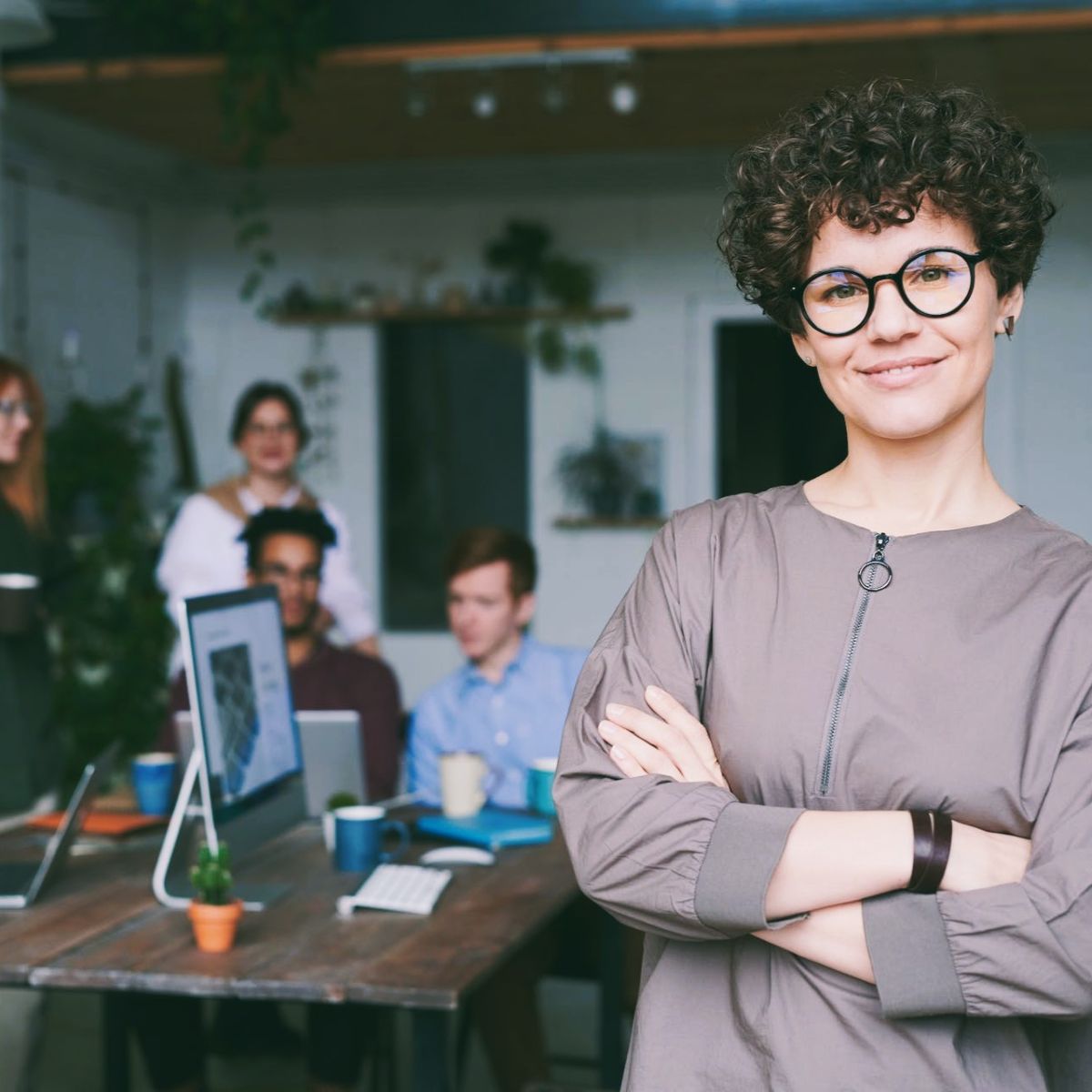 woman standing at forefront of camera with people meeting behind her in office setting.