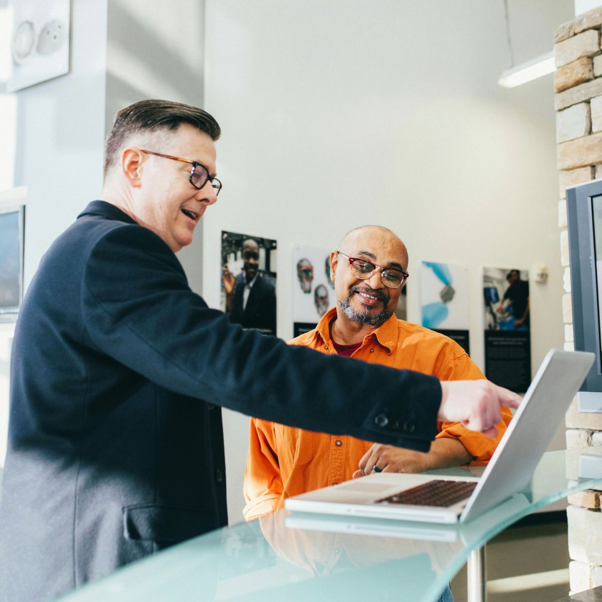 Man in suit talking to other man in orange shirt while pointing at computer.