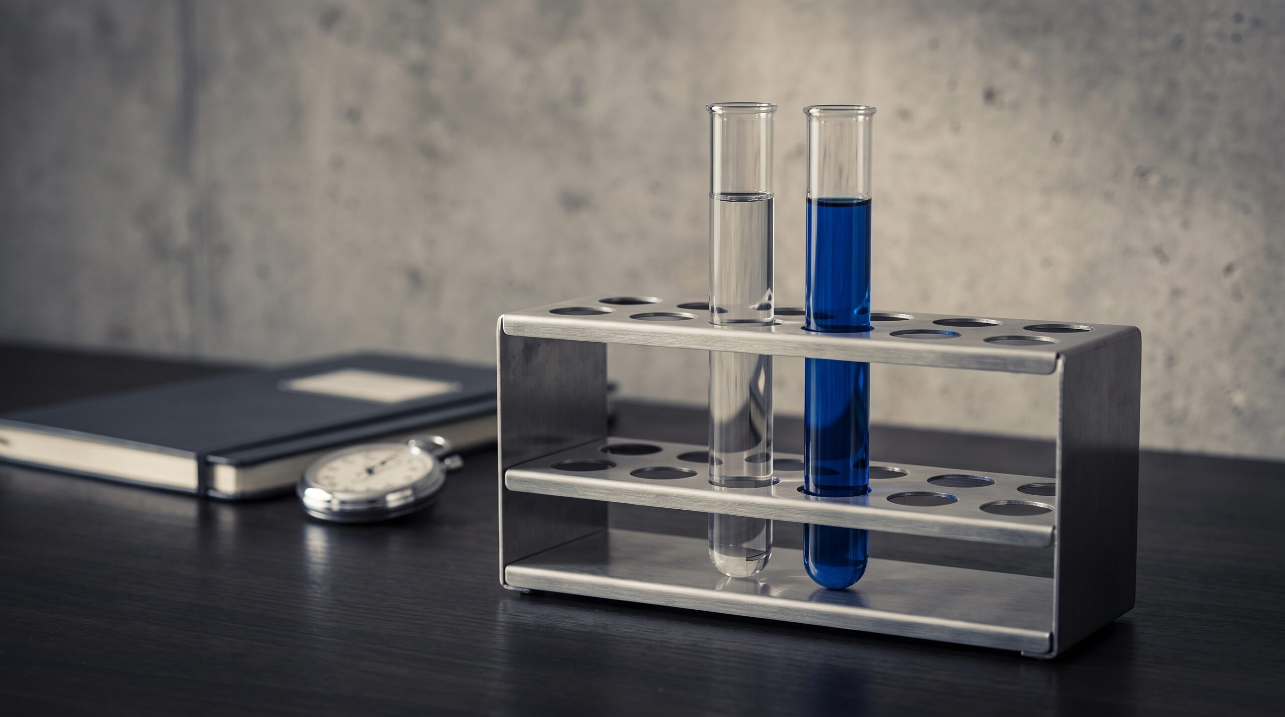 Two test tubes in a steel rack, one clear and one filled blue, beside a notebook and stopwatch: a control and a variant